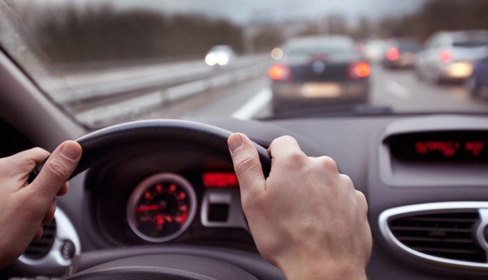 A view of the driver's side dashboard in a car from the driver's perspective with 2 hands on the wheel and cars in lanes viewed through the windshield.