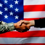 cropped view of soldier shaking hand with civilian man near american flag on blurred background