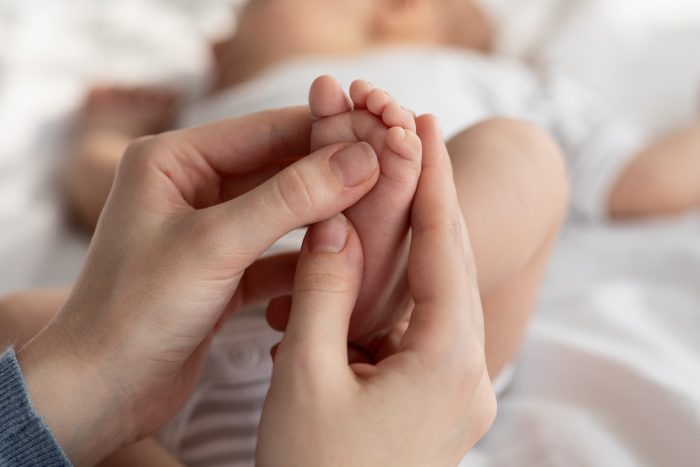 Closeup Shot Of Caring Mother Massaging Her Newborn Baby's Foot, Unrecognizable Loving Mom Making Strengthening Gymnastic Exercises For Her Sleeping Infant Child, Cropped Image, Selective Focus