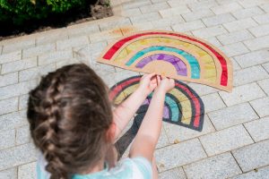 A little girl holding a rainbow suncatcher made of paper and tissue paper.