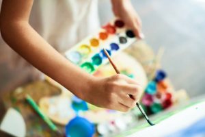 A picture of a kid's arms holding a watercolor palette in one hand and a paint brush touching a canvas in the other hand.