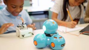 Diverse excited private STEM elementary school girls watch as their robots roll across the table. An Asian student records something in a notebook. An African American student works with the robots. Their mid adult Caucasian teacher helps them with the robots. Focus is on a blue robot in the foreground. The people are blurred in the background.