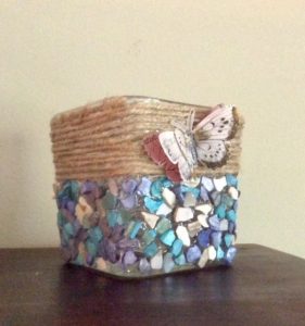 A votive candle holder wrapped with twine on the top half and various colored tile pieces on the bottom with a butterfly in the middle.
