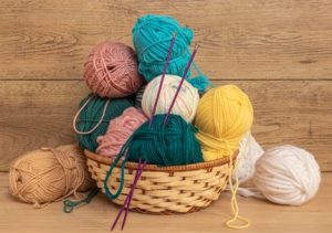 Different colored ball of yarn in a basket with a pair of knitting needles sticking out of the top of one.