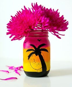 A mason jar painted pink, orange, yellow, and black to mimic a sunset with a palm tree.