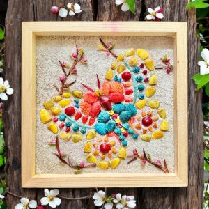 A butterfly made of various colored shells embedded in a sand bed and framed.