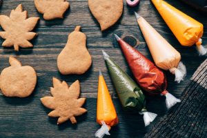 Cookies in the shapes of pears, pumpkins, and leaves on a table with bags of icing in yellow, orange, green, and red.