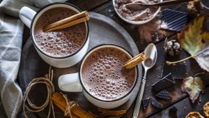 Two hot chocolate mugs shot from above on rustic wooden table. Some cinnamon sticks are on the table and inside the mugs and chocolate pieces are beside the mugs placed directly on the table. Low key DSRL studio photo taken with Canon EOS 5D Mk II and Canon EF 100mm f/2.8L Macro IS USM