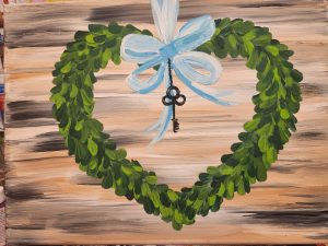 A green heart wreath with a key tied to a blue ribbon hanging down in the middle set against a wood-like background.