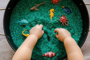 Toddler hands in a large bowl with a 1-2 inch layer of rice and different types of plastic sea creatures scattered throughout.