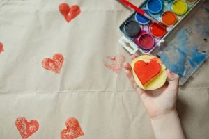 Child holds a potato stamp heart with red paint. Top view. Watercolor paint box in the background.