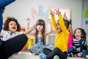 A small group of Kindergarten children sit on the floor of their classroom as their teacher lead them in a sing-along.  They are each dressed comfortably and are smiling as they do the actions to the song.